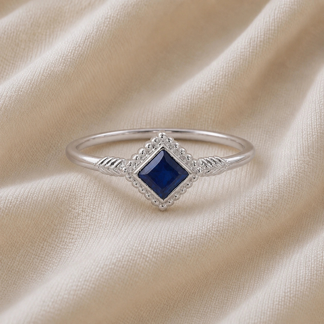 Princess_cut_ring_sterling_silver_with_square_sapphire_and_detailed_band_front_angle_view_on_soft_fabric_background