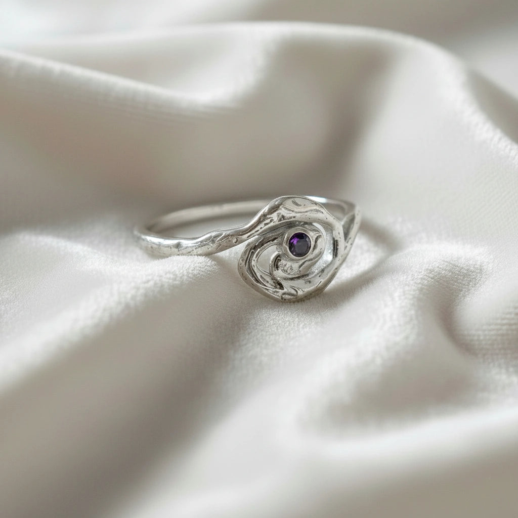 A side angle view of a silver amethyst ring with celestial jewelry swirl designs on an off white fabric background