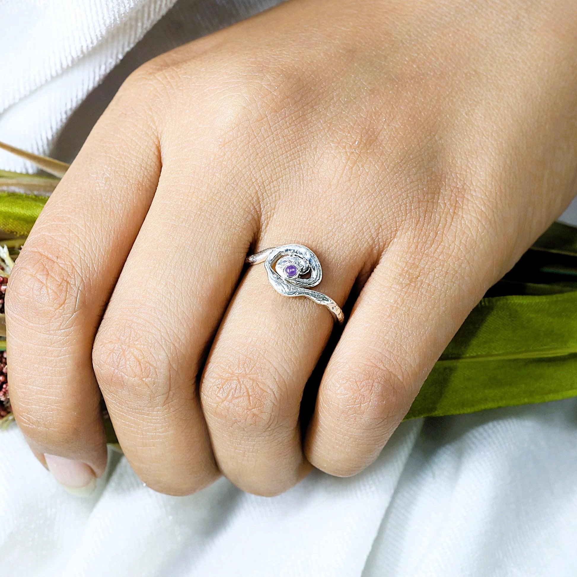 A silver amethyst ring with a textured swirl worn on a finger at a top angle over a white fabric background celestial jewelry