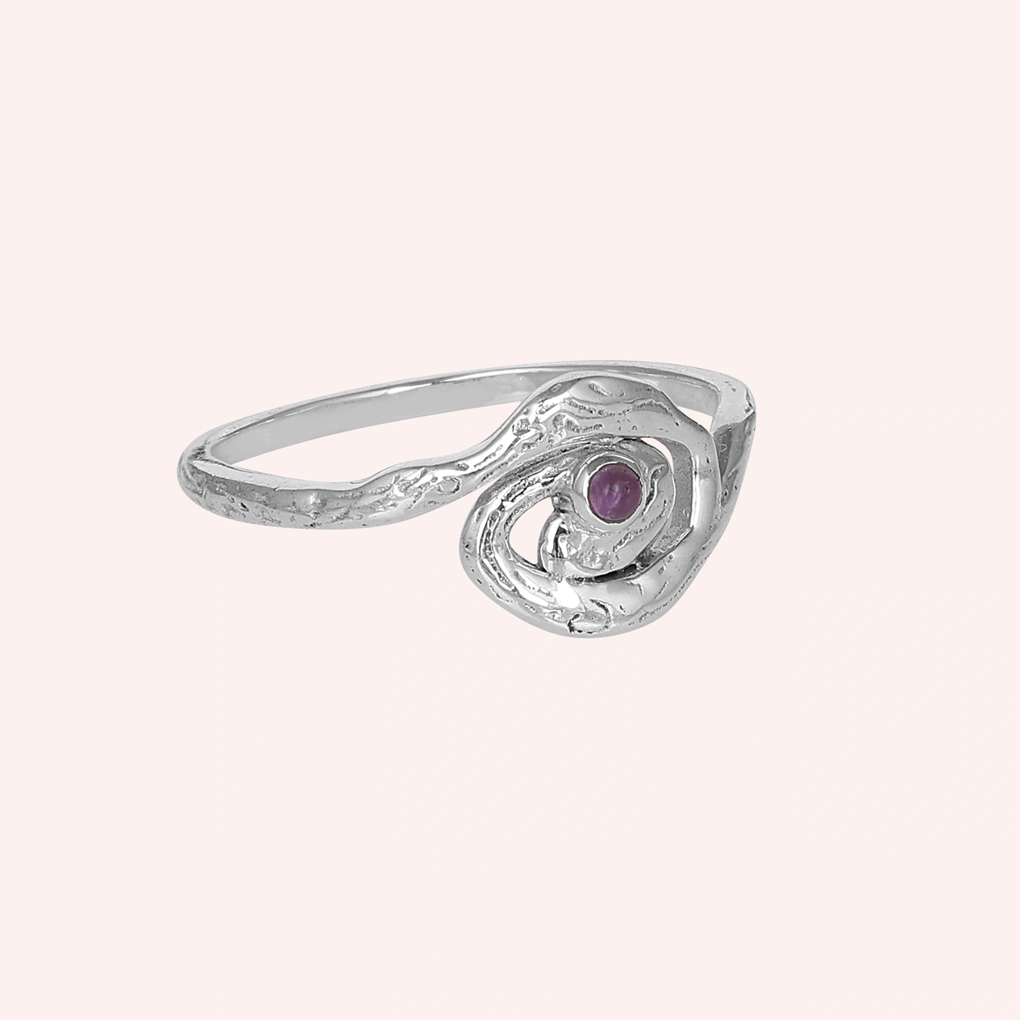 A silver amethyst ring with a swirl design shown from a side angle over a light pink background celestial jewelry inspired