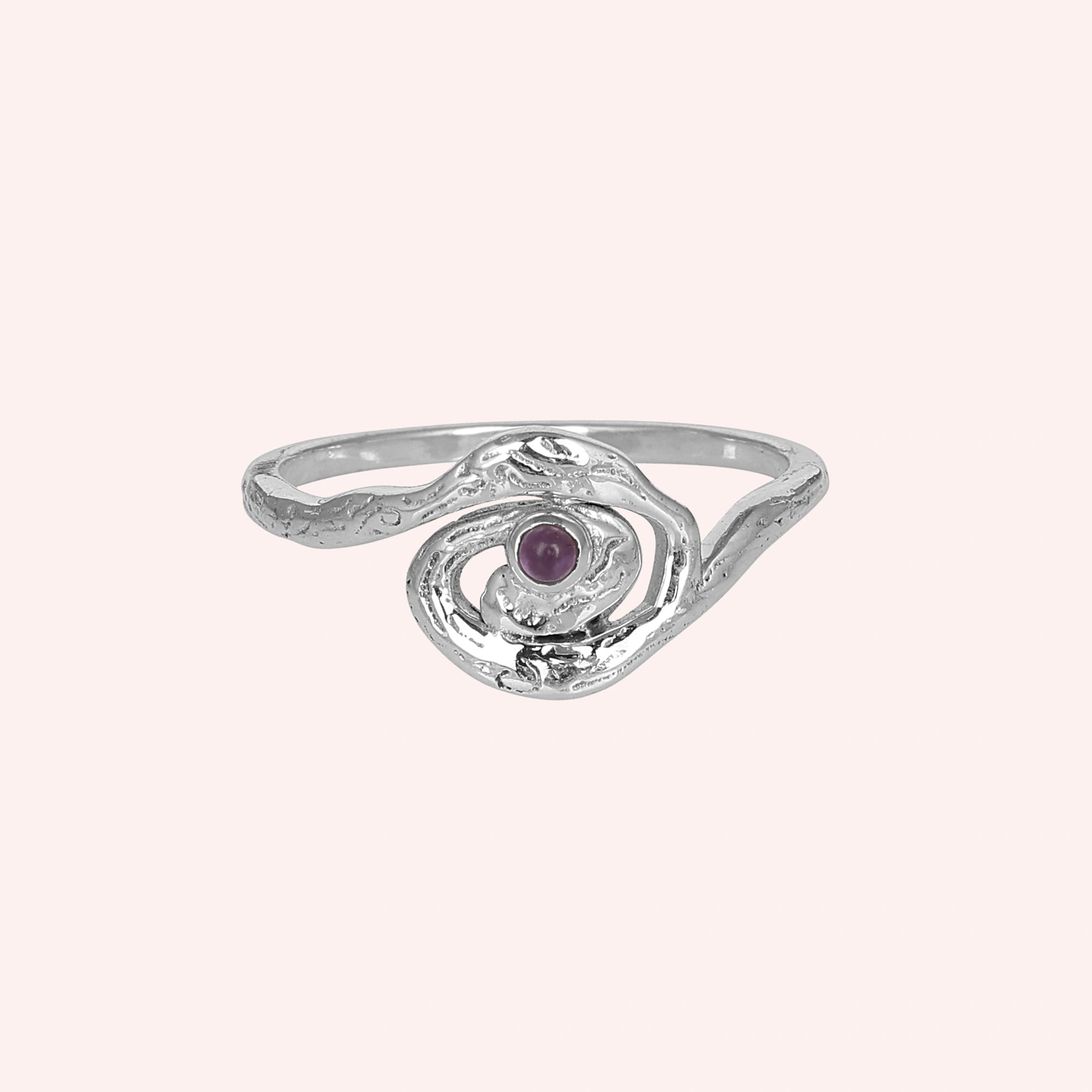 A front angle view of a silver amethyst ring with celestial jewelry swirl designs on a plain light pink solid color background