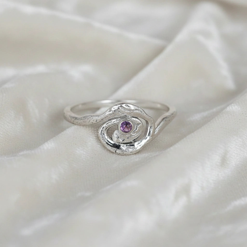 A front angle view of a silver amethyst ring with celestial jewelry swirl designs on an off white fabric background