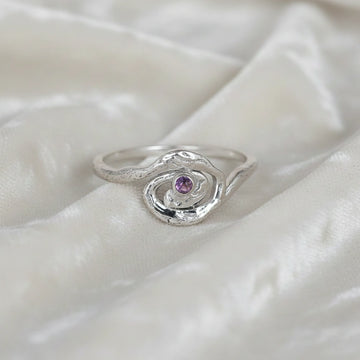 A front angle view of a silver amethyst ring with celestial jewelry swirl designs on an off white fabric background