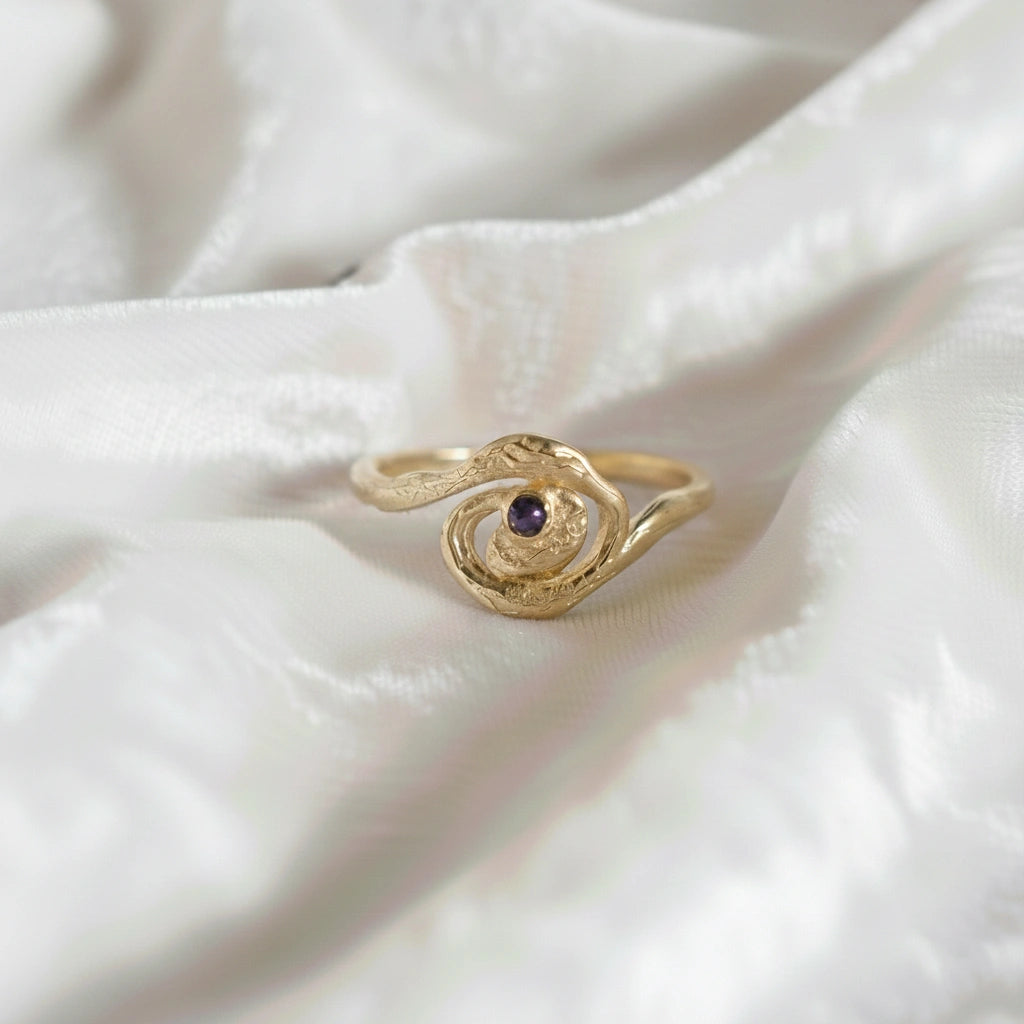 A gold plated silver amethyst ring with a galaxy swirl design at a front angle on an off white fabric background celestial jewelry