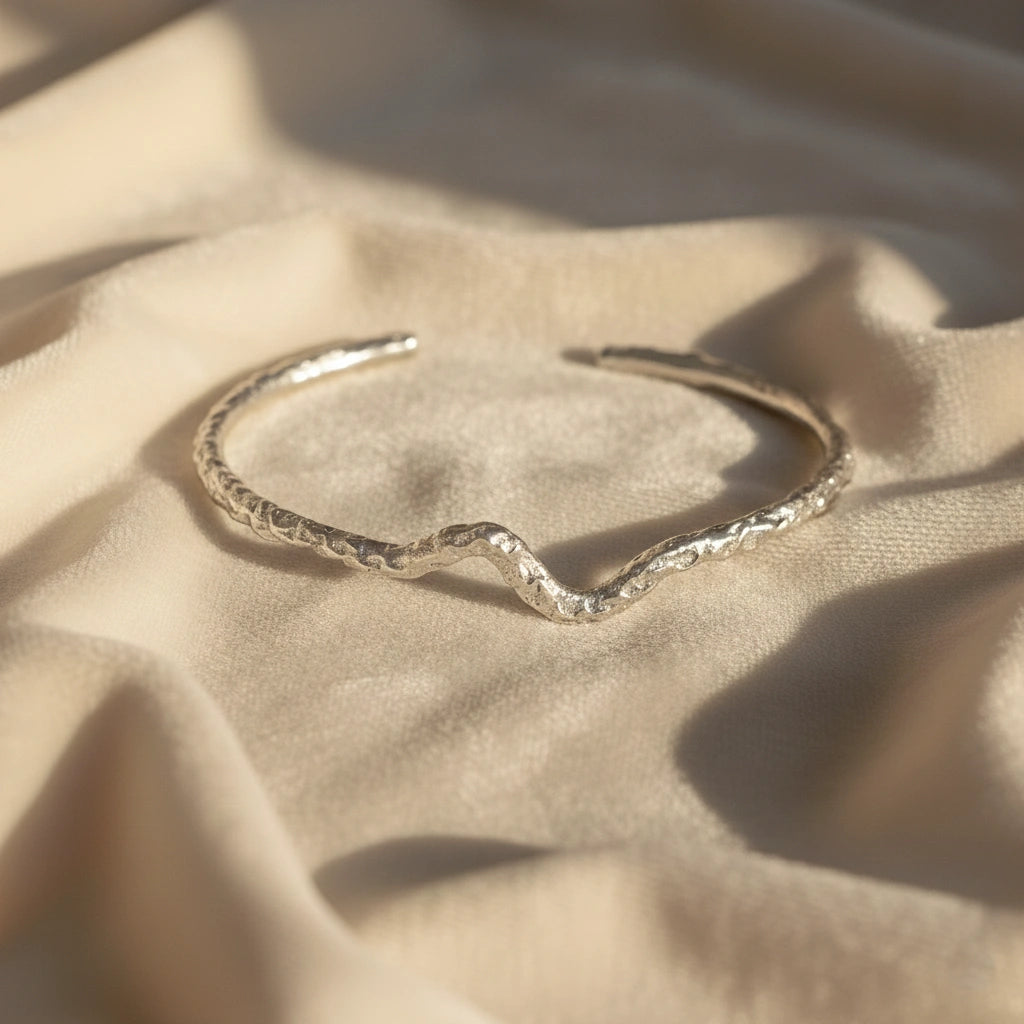 Silver cuff bracelet shown at front angled view featuring textured open silver design resting on soft beige fabric background
