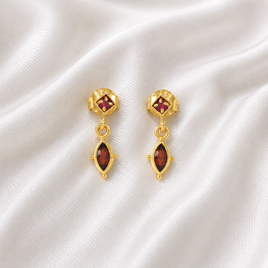 Silver_drop_earrings_in_gold_plated_silver_with_ruby_and_garnet_stones_at_front_angle_view_on_soft_fabric_background