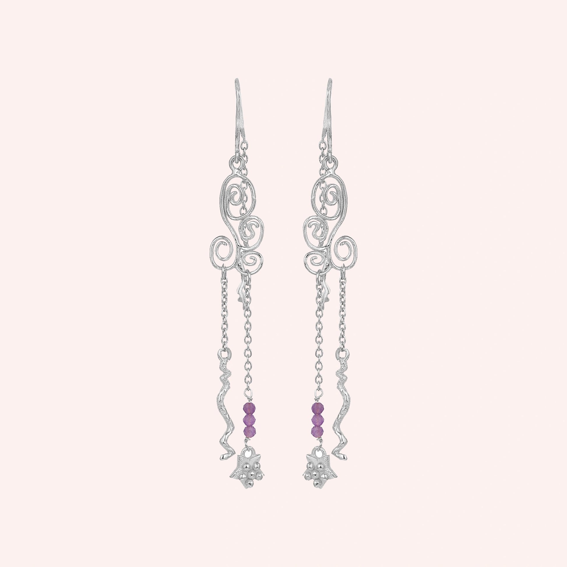 front_angle_silver_hanging_earrings_with_scroll_design_and_purple_beads_showing_trendy_earrings_on_soft_pink_background