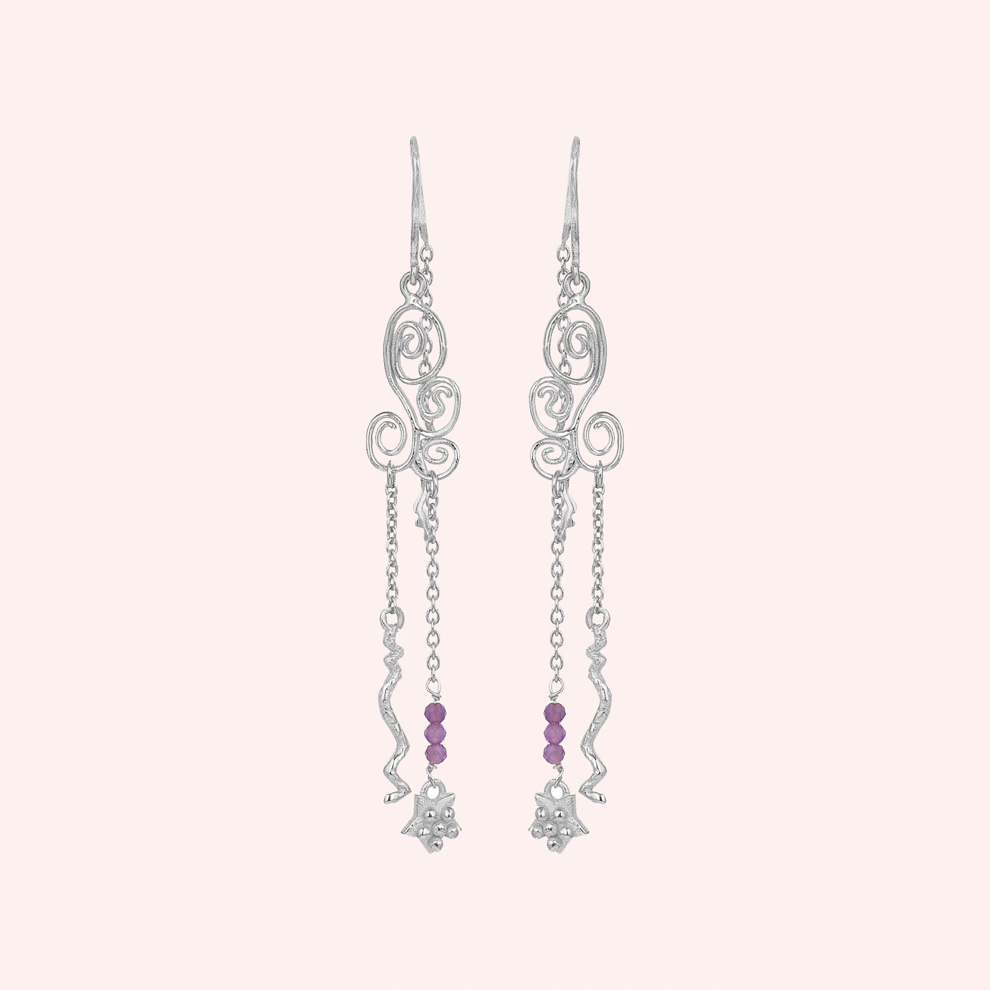 front_angle_silver_hanging_earrings_with_scroll_design_and_purple_beads_showing_trendy_earrings_on_soft_pink_background