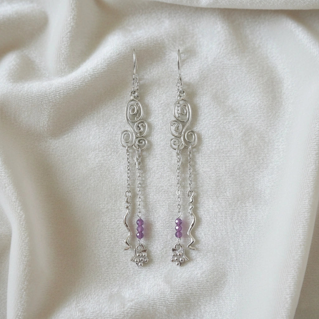 front_angle_silver_hanging_earrings_with_scroll_design_purple_beads_and_chain_details_trendy_earrings_on_soft_white_fabric