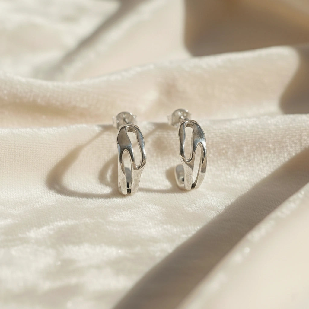 Silver hoop earrings shown at front angled view featuring sculpted open hoop design in polished silver on a soft fabric background