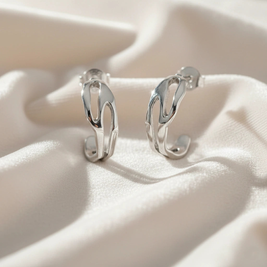 Silver hoop earrings shown at front angled view featuring sculpted open hoop design in silver resting on soft fabric background