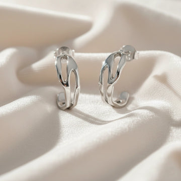 Silver hoop earrings shown at front angled view featuring sculpted open hoop design in silver resting on soft fabric background