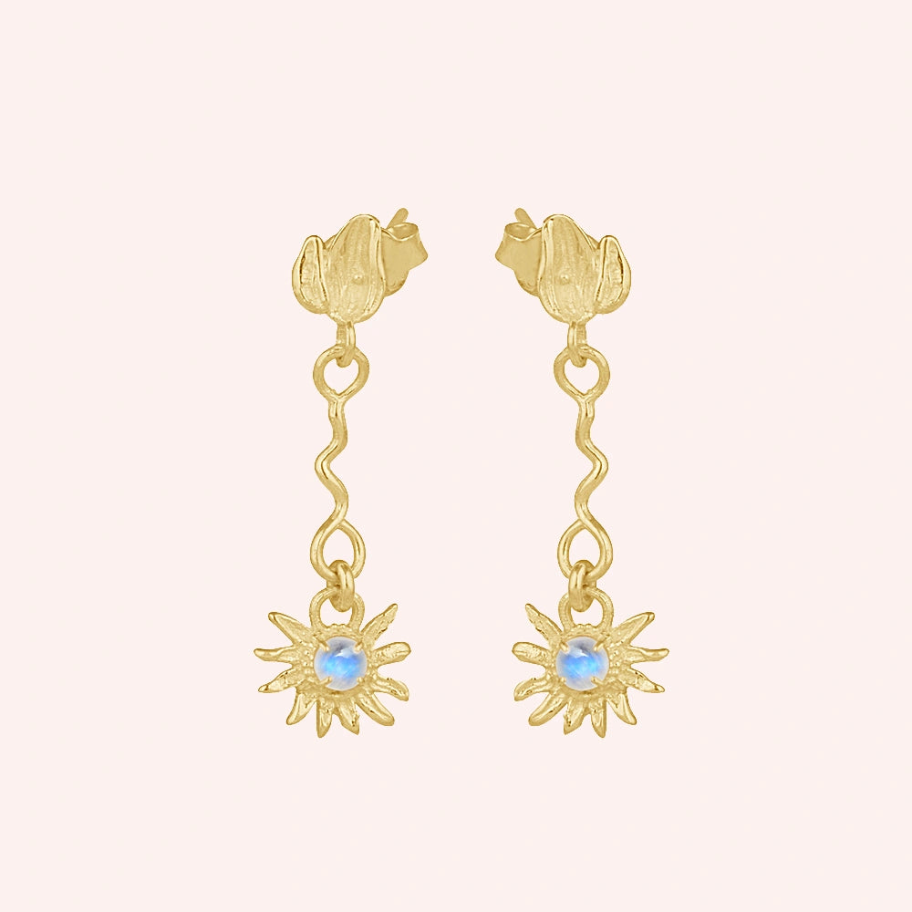 front angle of gold plated moonstone earrings and flower earrings featuring a wavy drop design on a plain pink background