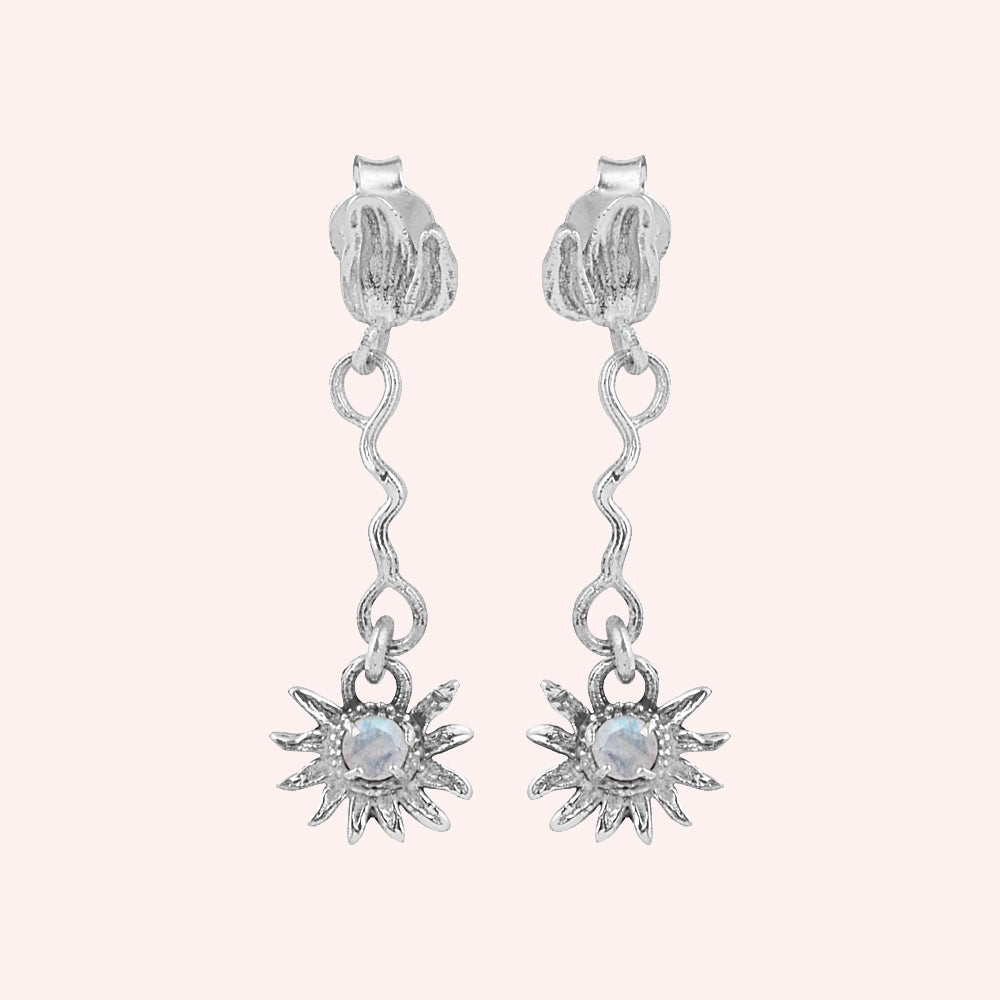 front angle of moonstone earrings and flower earrings in silver featuring a wavy drop design on a plain pink background