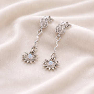 a close up high angle shot of moonstone earrings and flower earrings in silver on a soft cream fabric with wavy textures