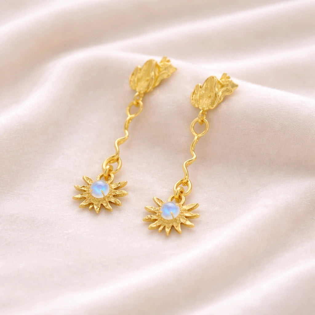 high angle of gold plated moonstone earrings and flower earrings with a wavy drop design resting on soft cream wavy fabric
