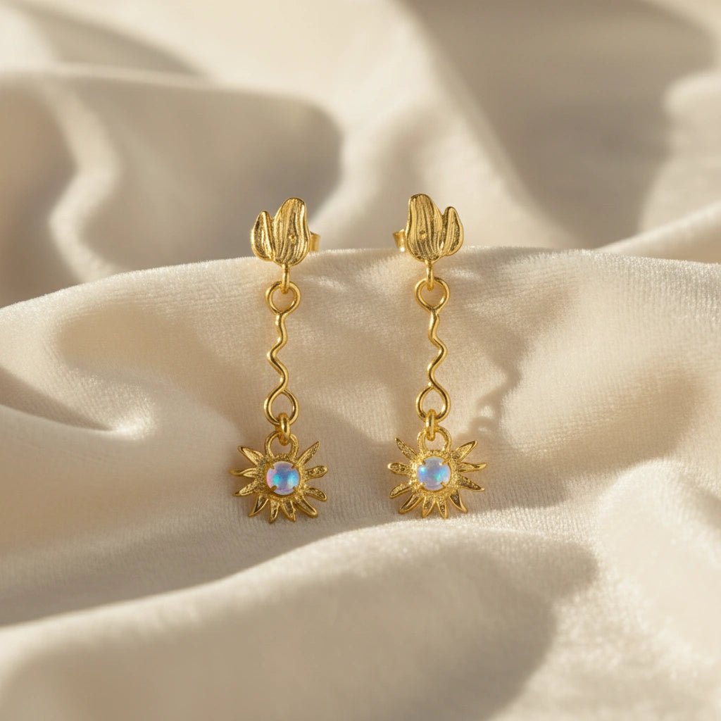 front angle of gold plated moonstone earrings and flower earrings with a wavy drop design resting on soft white wavy fabric