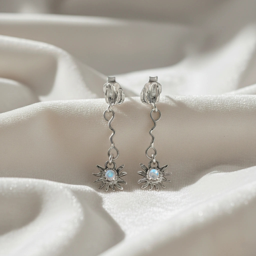front angle of silver moonstone earrings and flower earrings with a wavy drop design resting on soft white wavy fabric texture