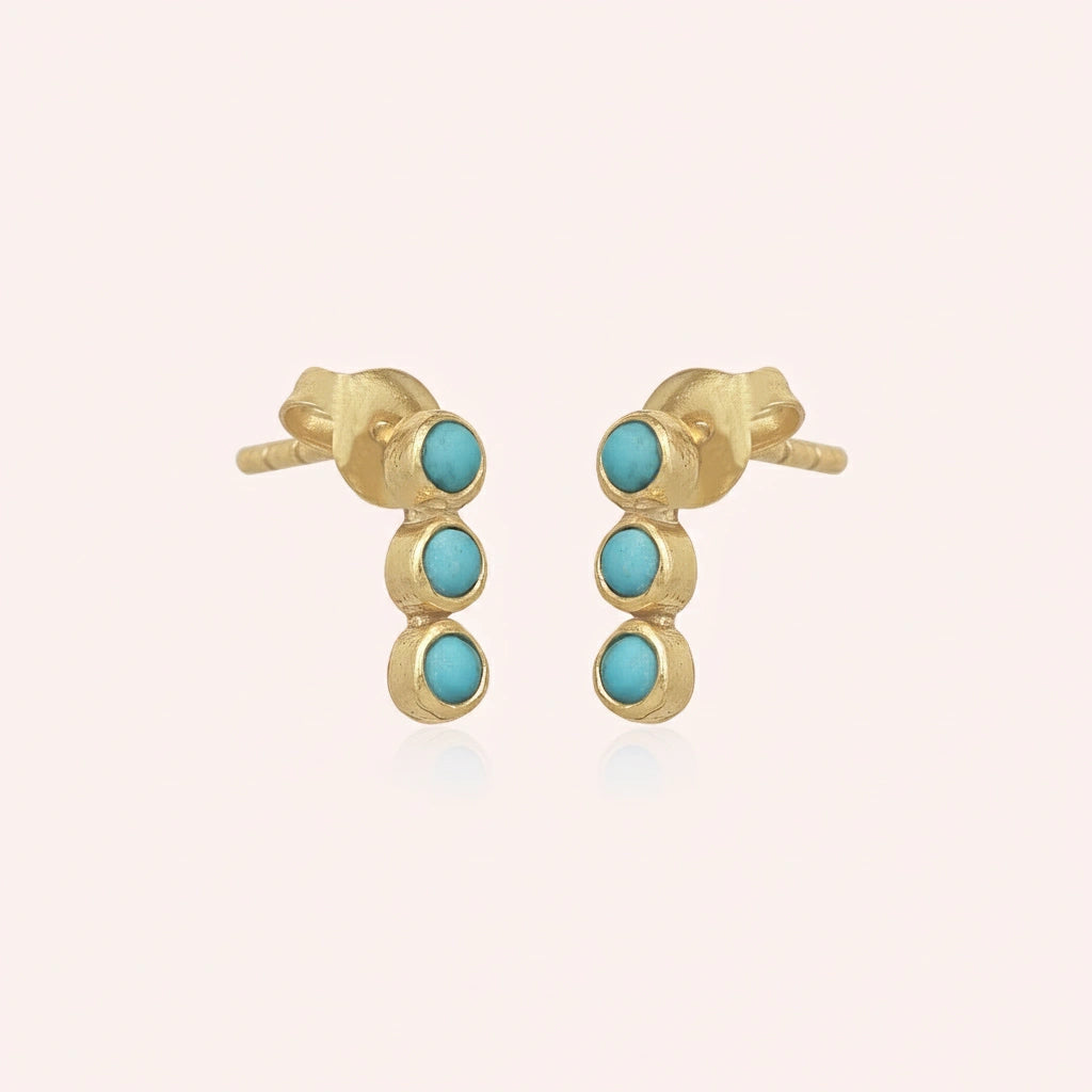 A gold plated silver stud earring set with turquoise earrings in a vertical line at a side angle over a pink background