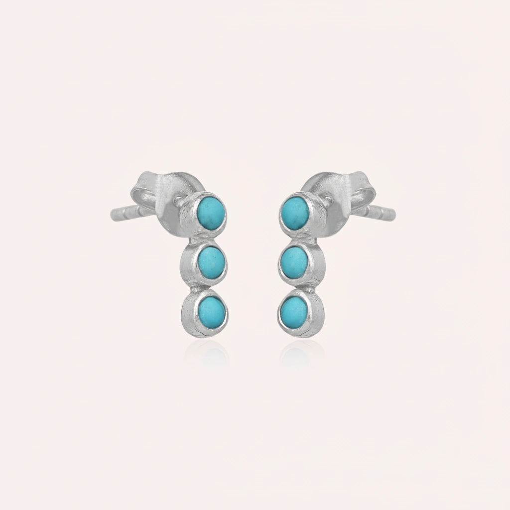 A pair of silver stud earrings with turquoise earrings stones in a vertical row at a side angle against a pink backdrop