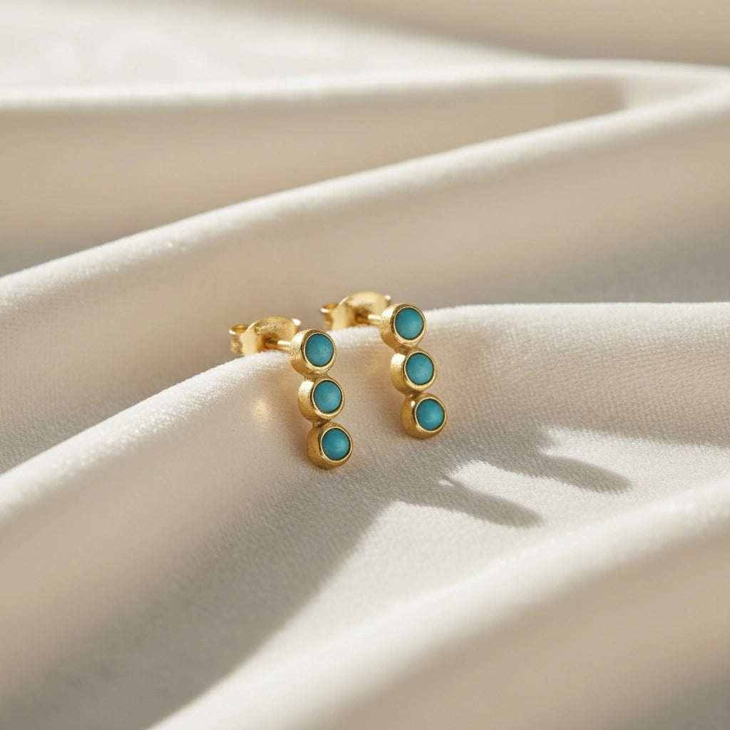Gold plated silver stud earrings with turquoise earrings stones set in a vertical line at a side angle on pink backdrop