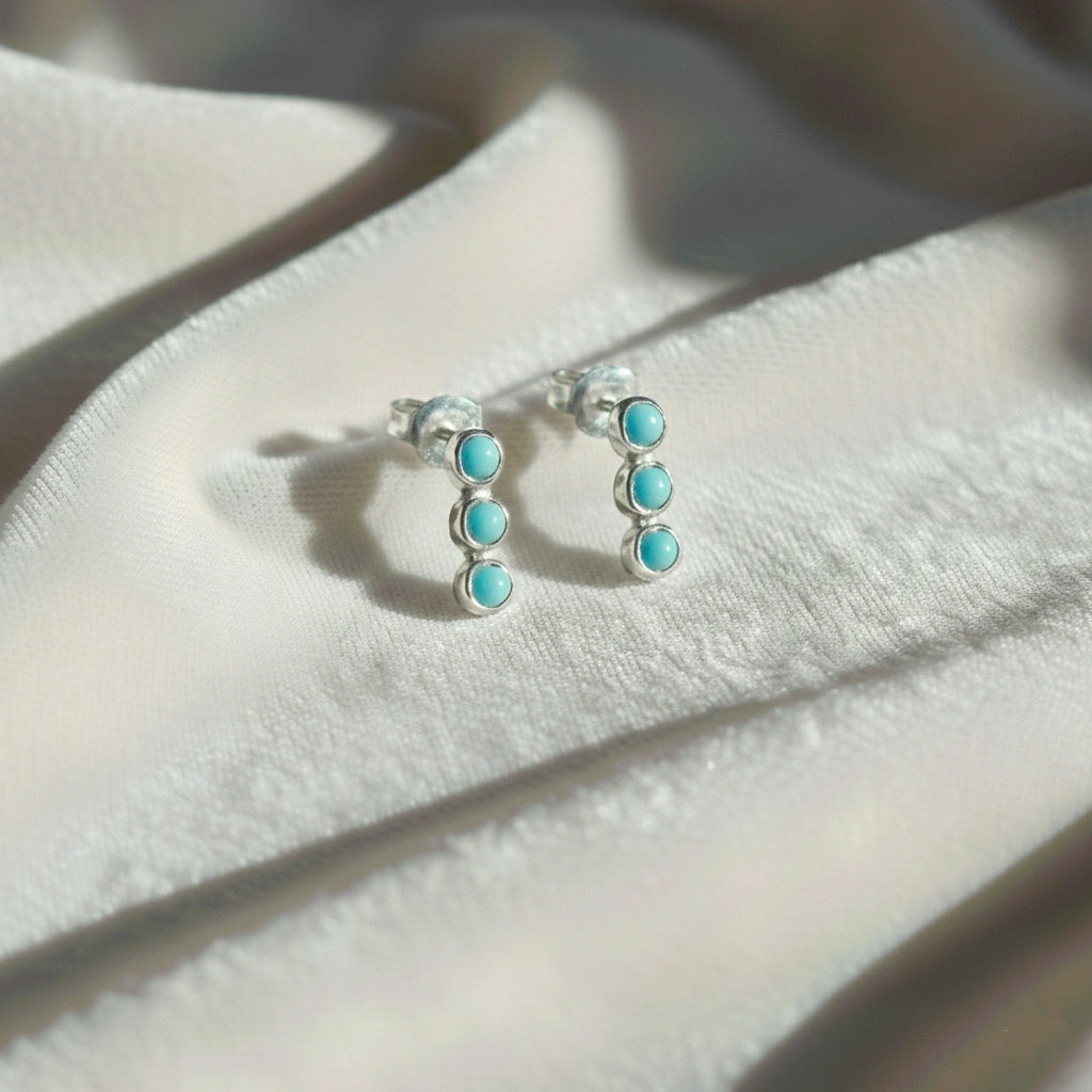Pair of silver stud earrings with turquoise earrings stones set in a vertical row at a side angle on white velvet fabric