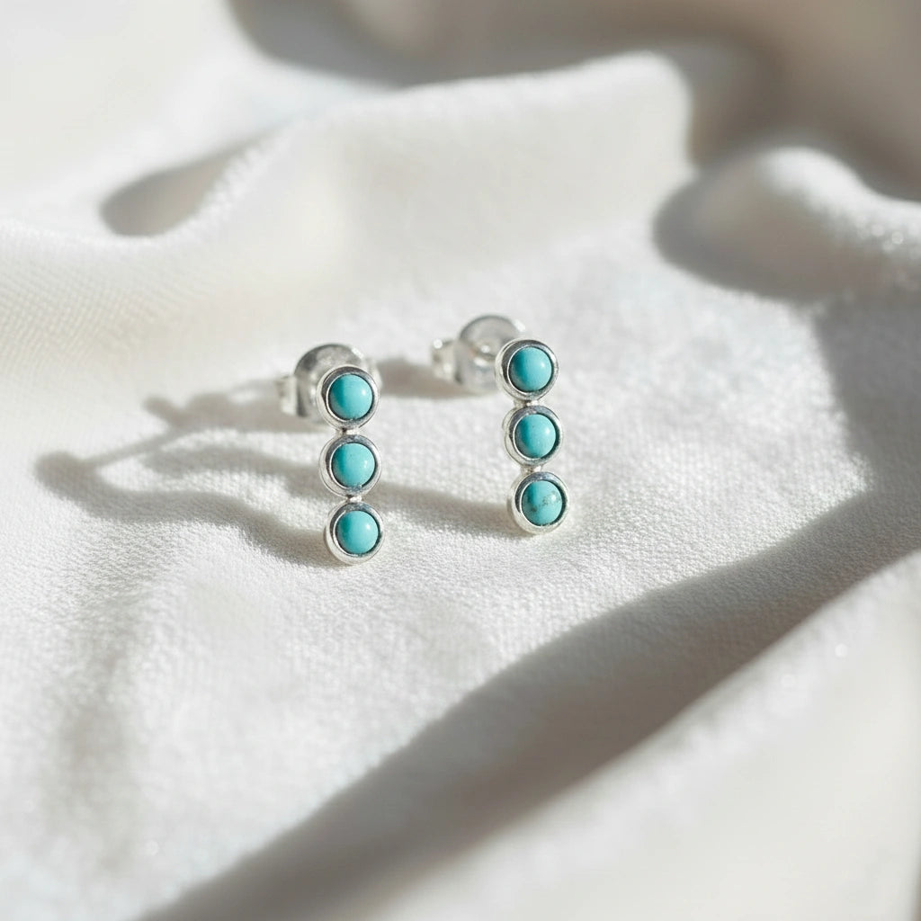 Pair of silver stud earrings with turquoise earrings stones set in a vertical row at a straight angle on white velvet fabric