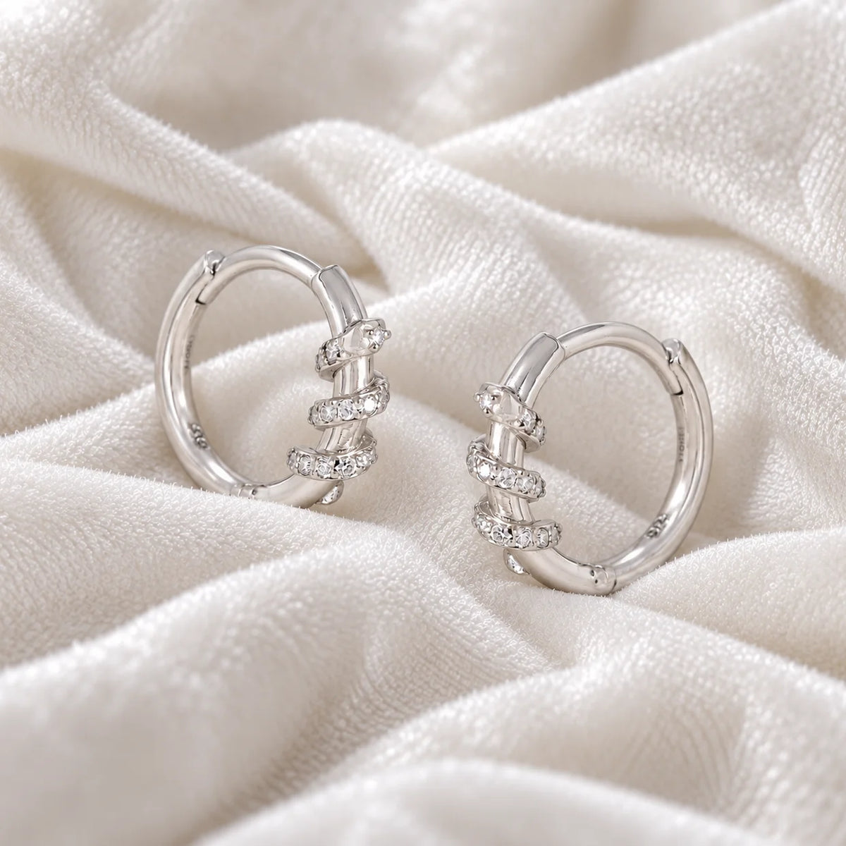 Small_hoop_earrings_in_sterling_silver_with_a_twisted_design_band_at_side_angle_view_on_soft_fabric_background