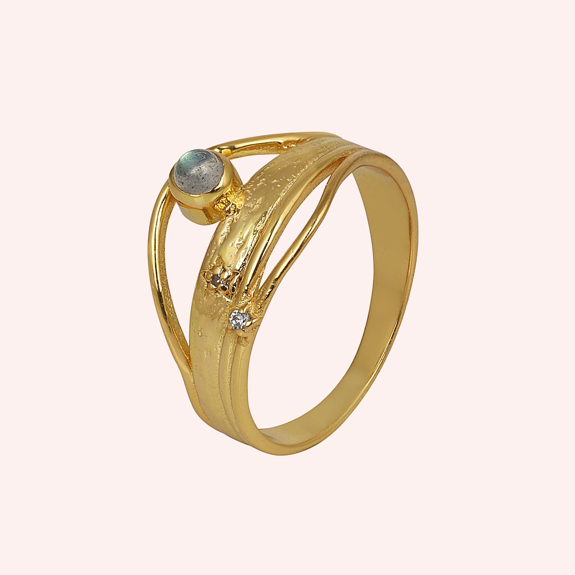 A gold plated wave ring with a labradorite on a pink background shown from a side angle as a textured statement ring design