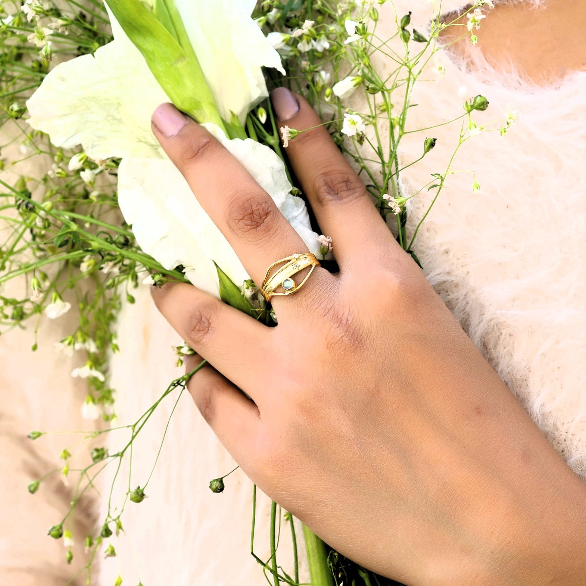A gold plated wave ring held at a top angle on a hand with flowers against a fuzzy sweater as a textured statement ring design