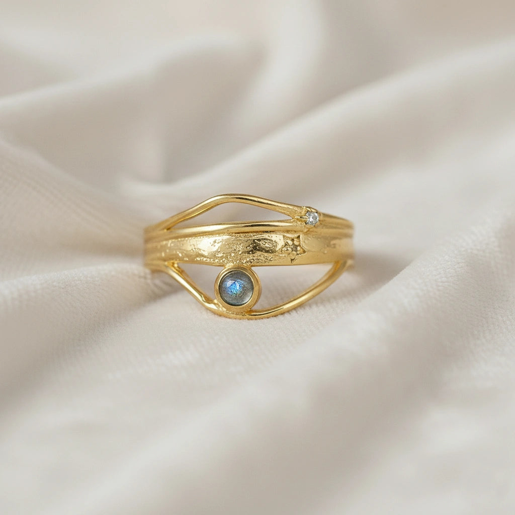 A gold plated wave ring with a labradorite on a white fabric shown from a front angle as a textured statement ring design