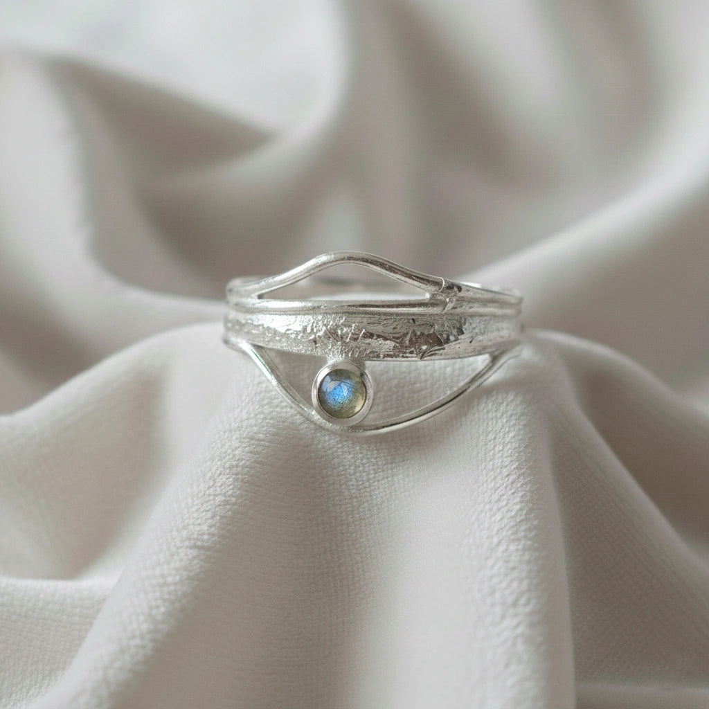 A silver wave ring statement ring with a textured band and round labradorite stone at a front angle at a white fabric background