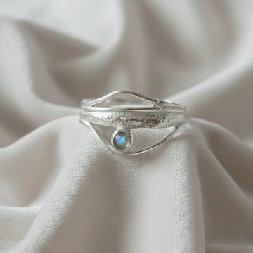 A silver wave ring statement ring with a textured band and round labradorite stone at a front angle at a white fabric background