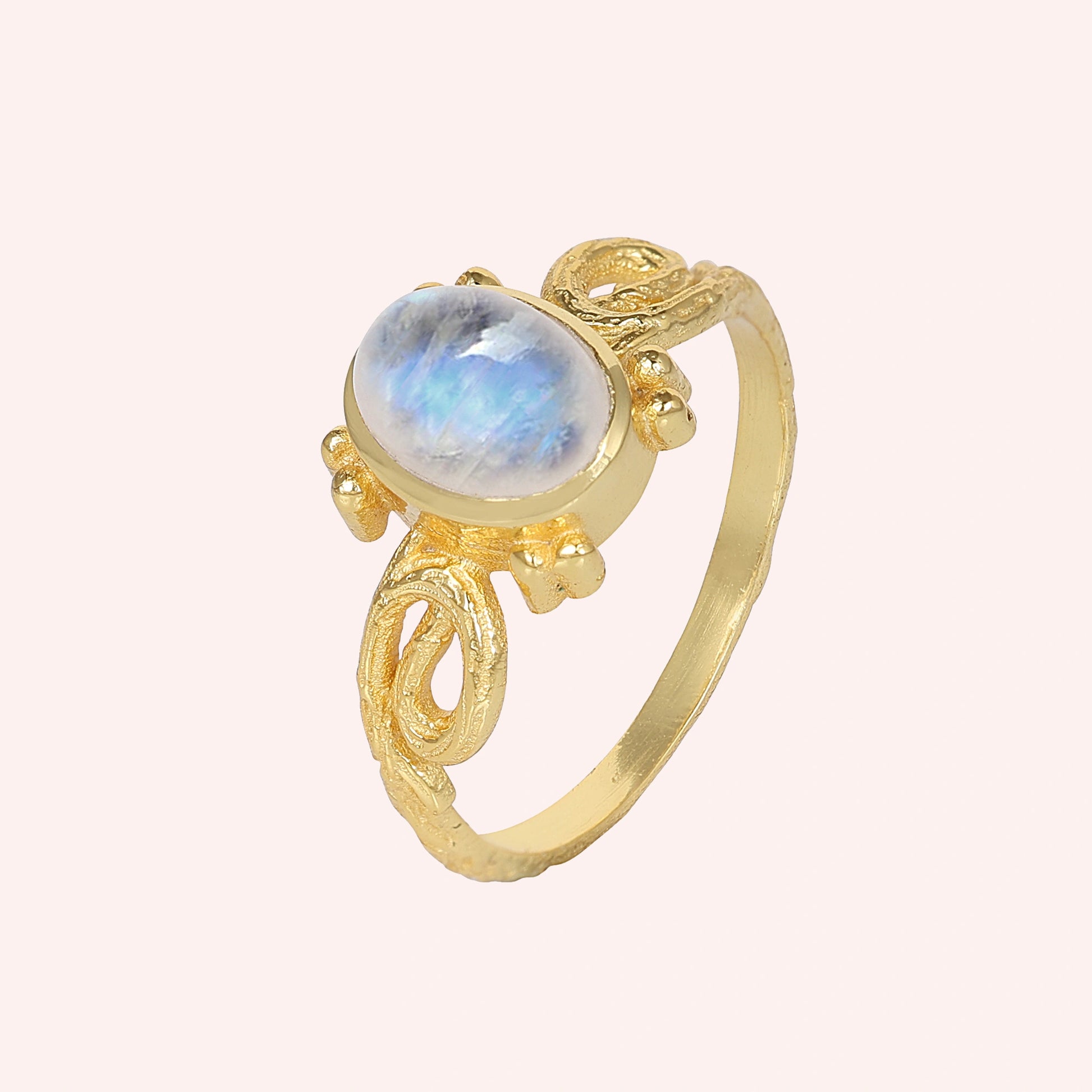 Statement_ring_gold_plated_silver_with_oval_moonstone_and_intricate_band_detail_slightly_tilted_view_on_solid_pink_background