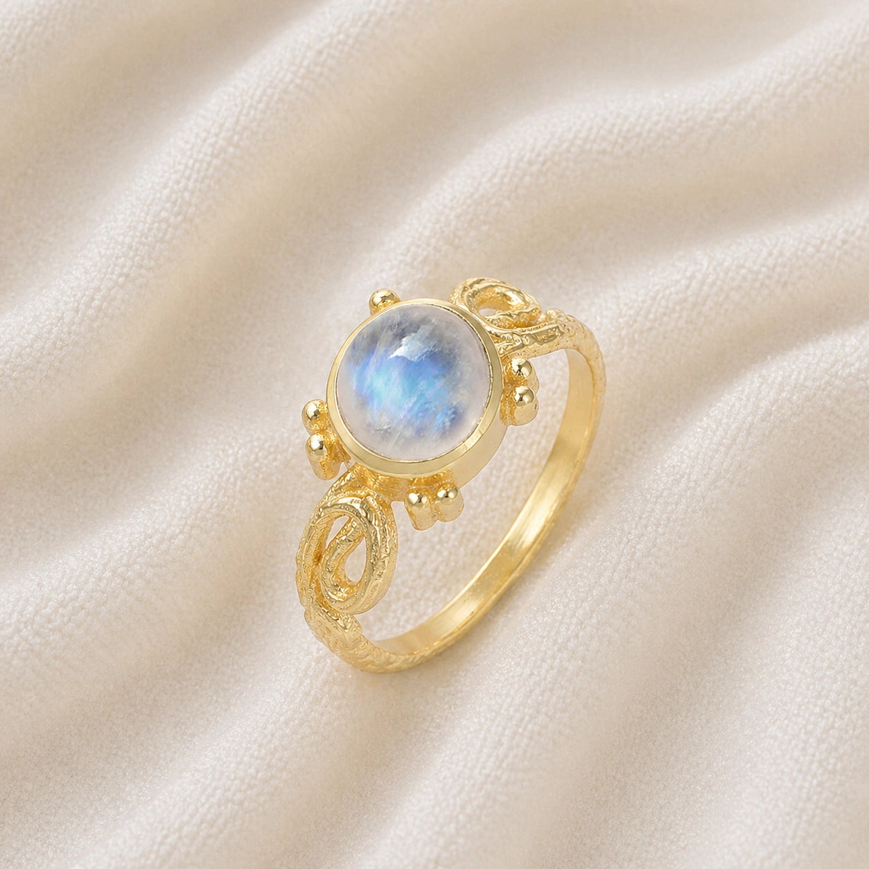 Statement_ring_gold_plated_silver_with_oval_moonstone_and_intricate_band_detail_slightly_tilted_view_on_soft_fabric_background