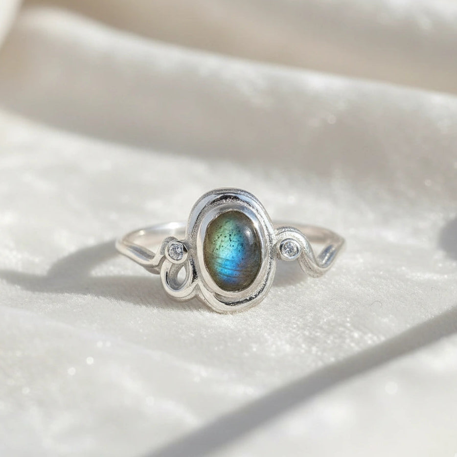 Front view of a sterling silver labradorite ring featuring an oval labradorite silver wavy ring band on white fabric background