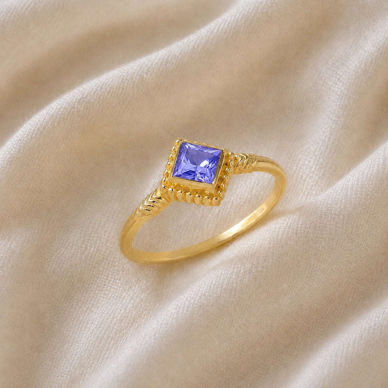 Princess_cut_ring_gold_plated_silver_with_square_tanzanite_and_detailed_band_slightly_tilted_view_on_soft_fabric_background