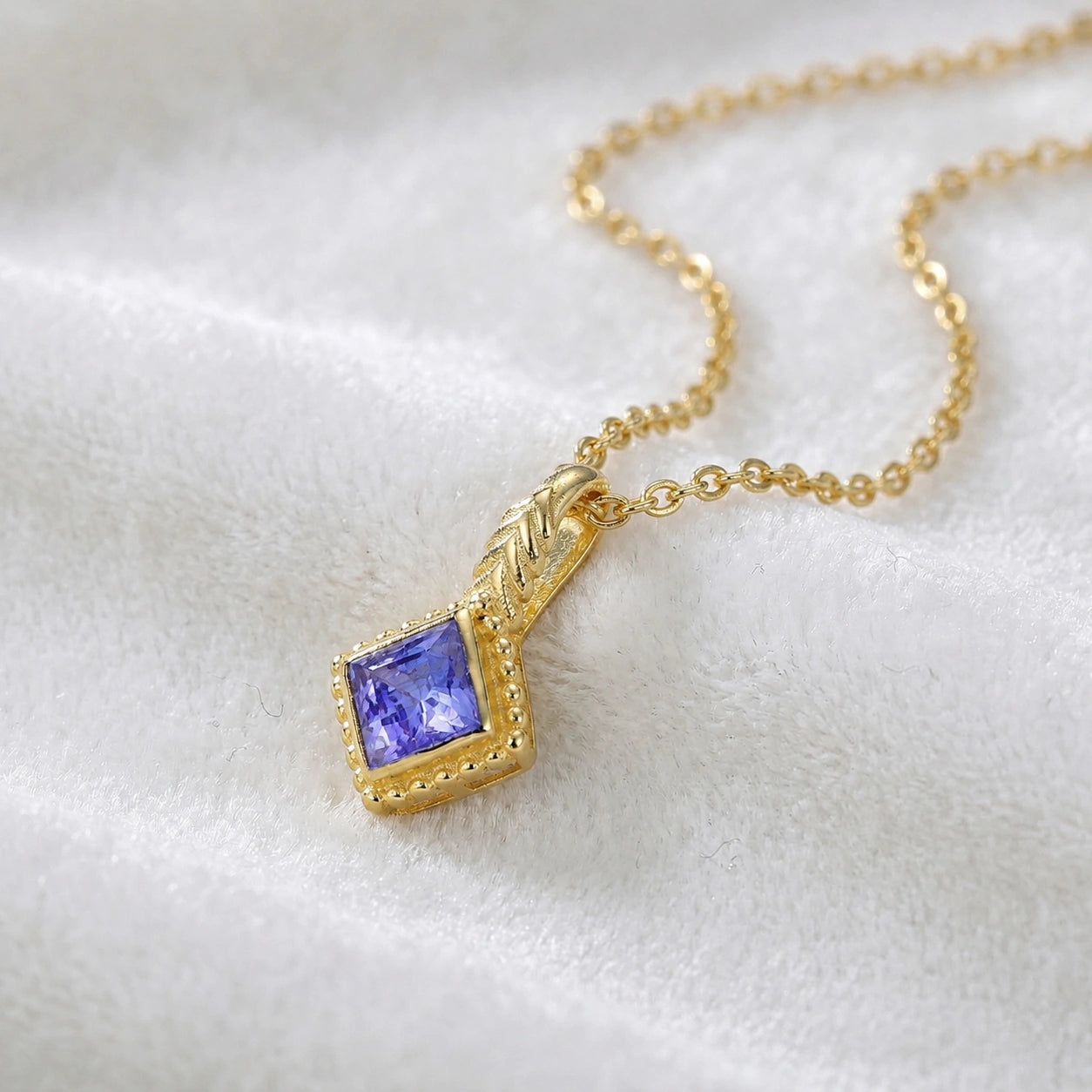 Birthstone_necklace_in_gold_plated_silver_with_square_cut_tanzanite_pendant_in_slightly_angled_view_on_soft_white_fabric_background