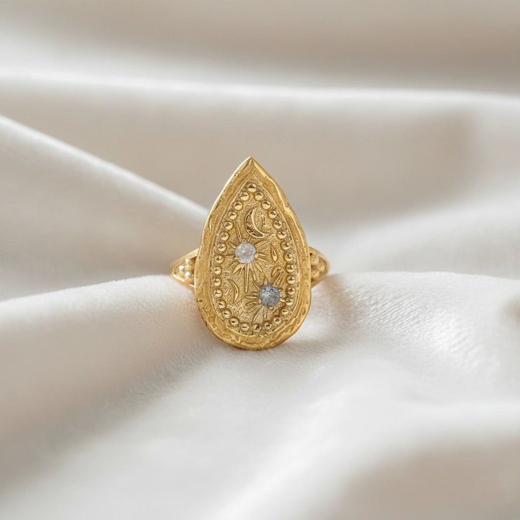 Gold plated celestial ring featuring a large teardrop design with carved sun moon and stars on a white cloth at a straight on angle