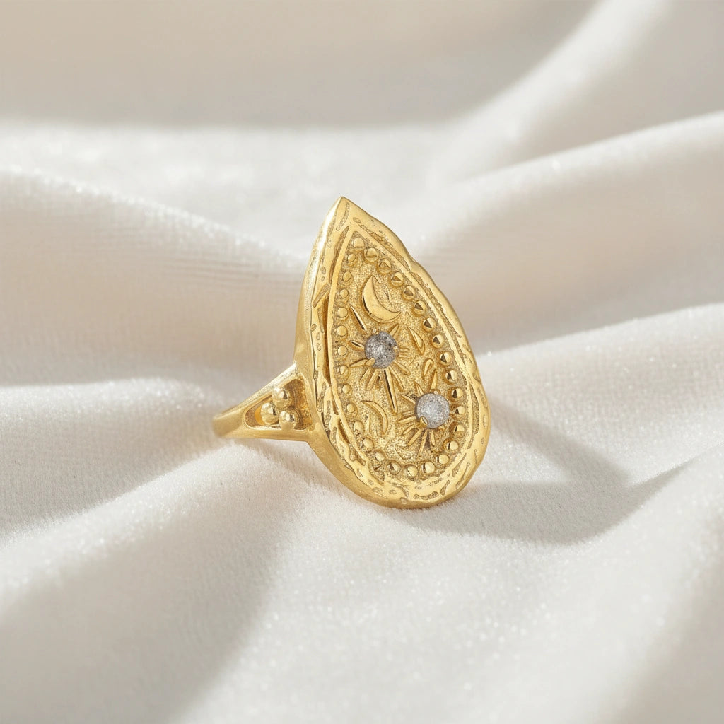 Gold plated celestial ring featuring a large teardrop design with carved sun moon and stars on a white cloth angled view