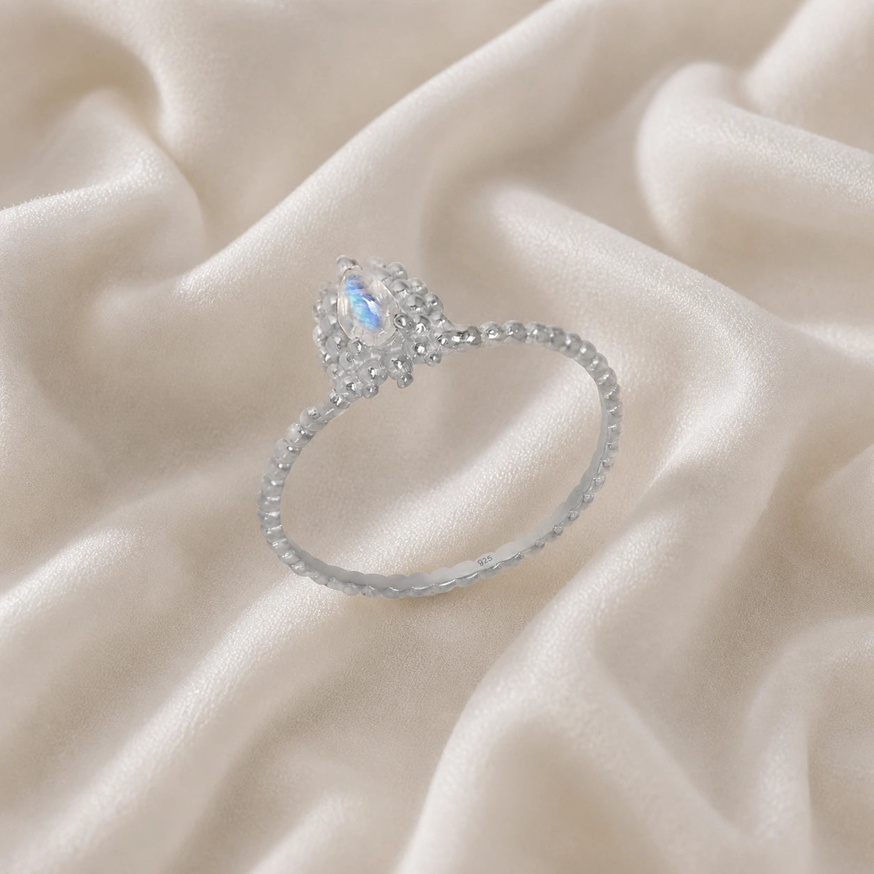 Teardrop_ring_sterling_silver_beaded_band_with_moonstone_at_tilted_angle_view_on_soft_fabric_background