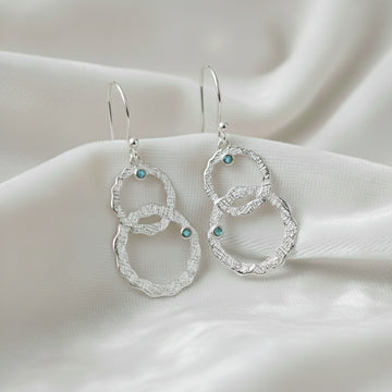 Silver dangle earrings textured interlocking rings with small labradorite stones viewed straight on a soft white draped fabric