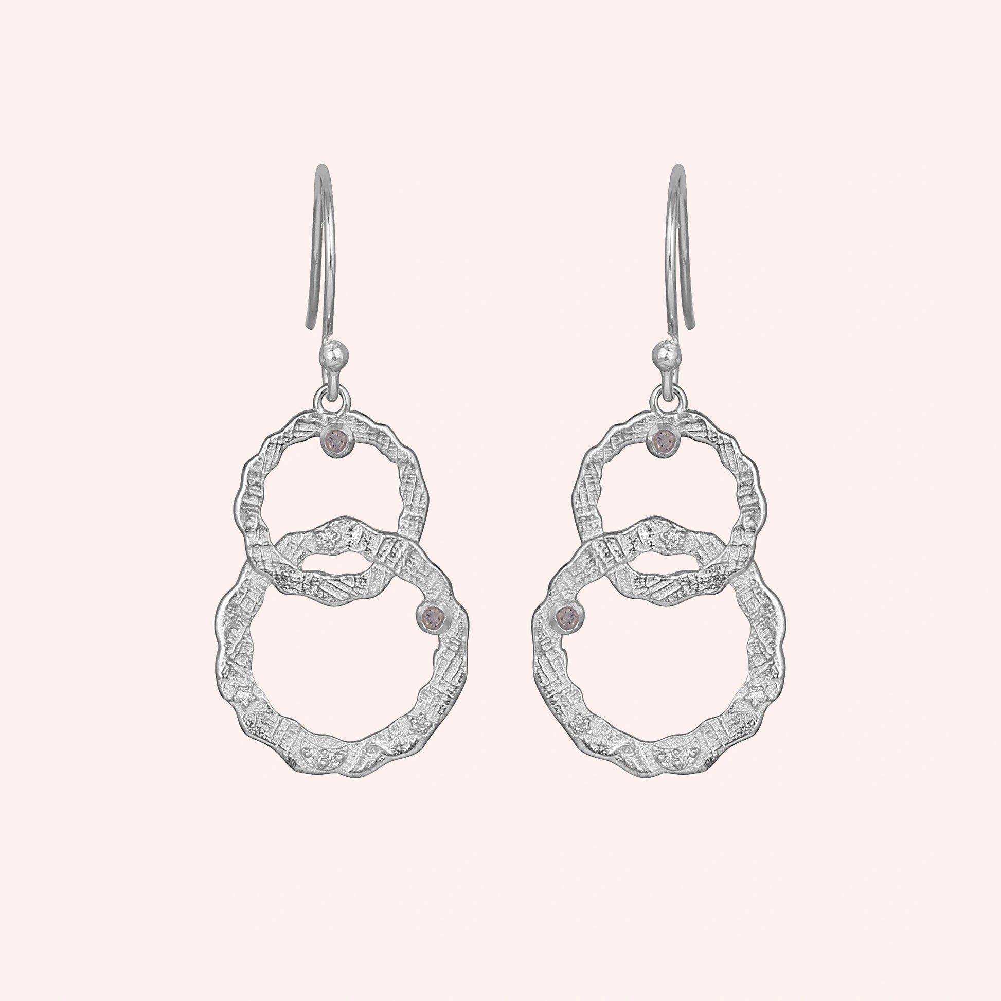 Silver dangle earrings textured interlocking rings and small labradorite stones viewed straight on a light pink background
