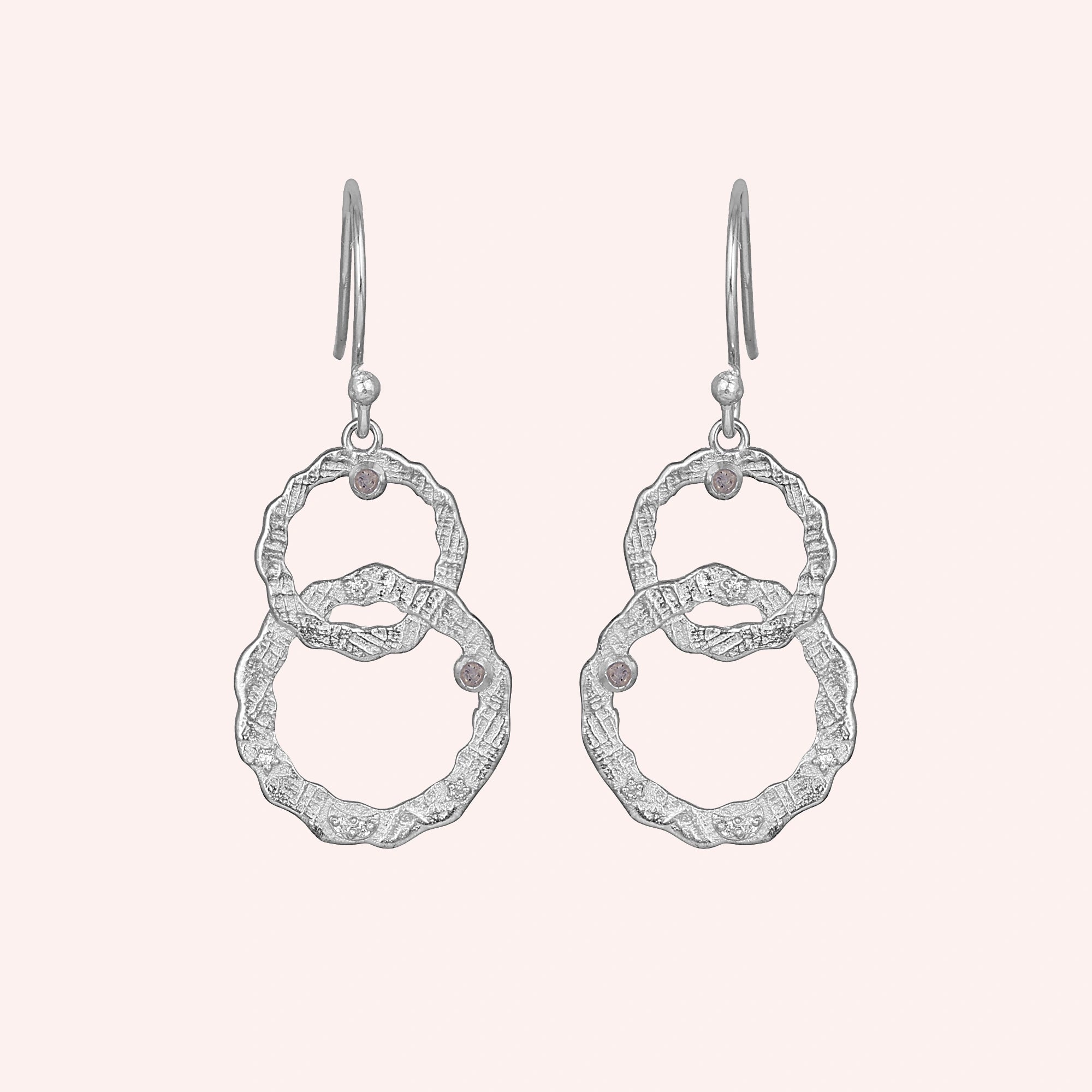 Silver dangle earrings textured interlocking rings and small labradorite stones viewed straight on a light pink background