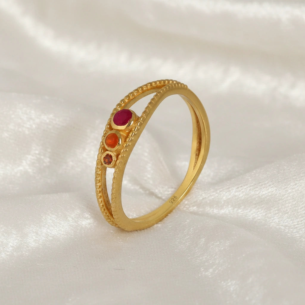 Three-stone-ring-in-gold-plated-silver-with-pink-red-and-orange-stones-in-angled-side-view-on-soft-white-fabric-background