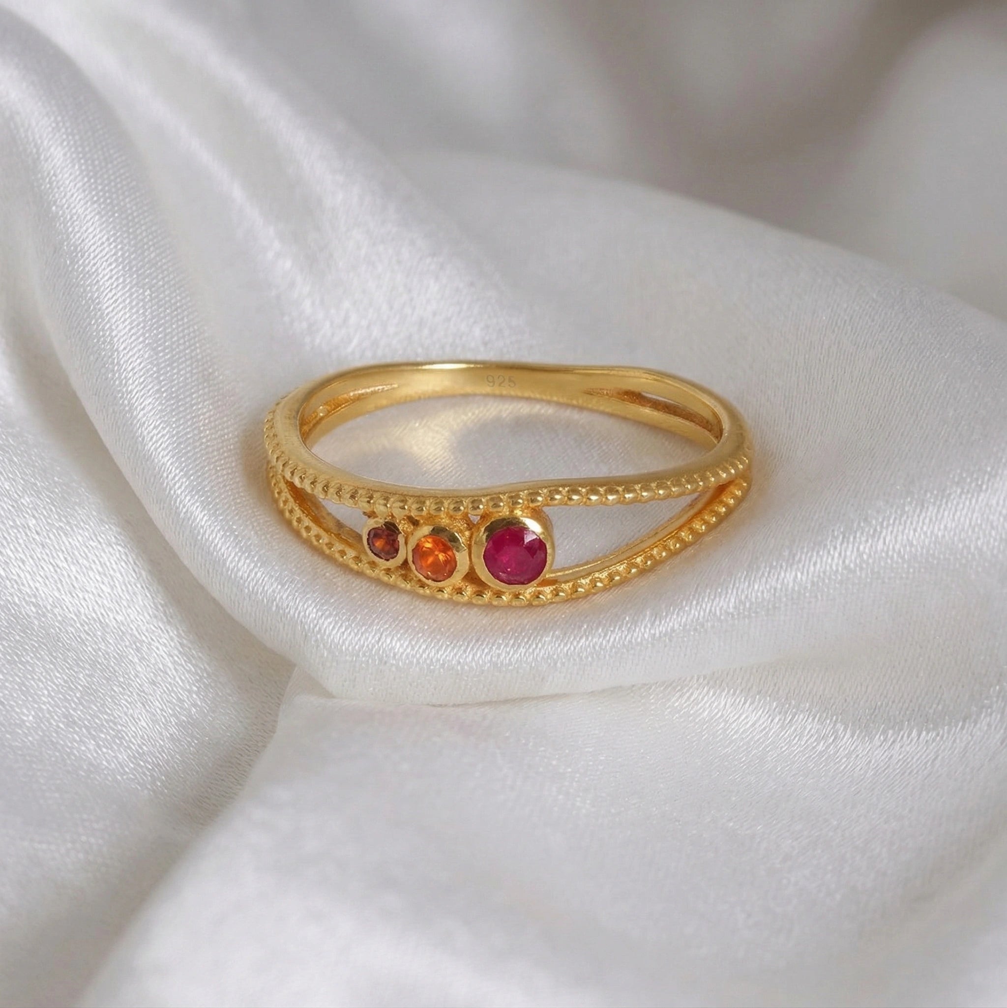 Three-stone-ring-in-gold-plated-silver-with-pink-red-and-orange-stones-in-front-view-on-soft-white-fabric-background