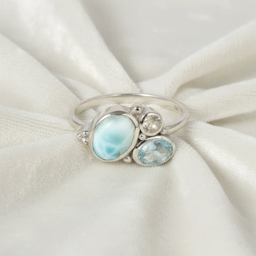 Detailed blue stone ring with Larimar Blue Topaz Moonstone resting on an off white draped fabric background viewed straight on