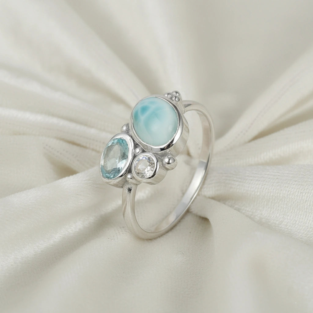 Detailed blue stone ring with Larimar Blue Topaz Moonstone resting on a soft white draped fabric background viewed from an angle