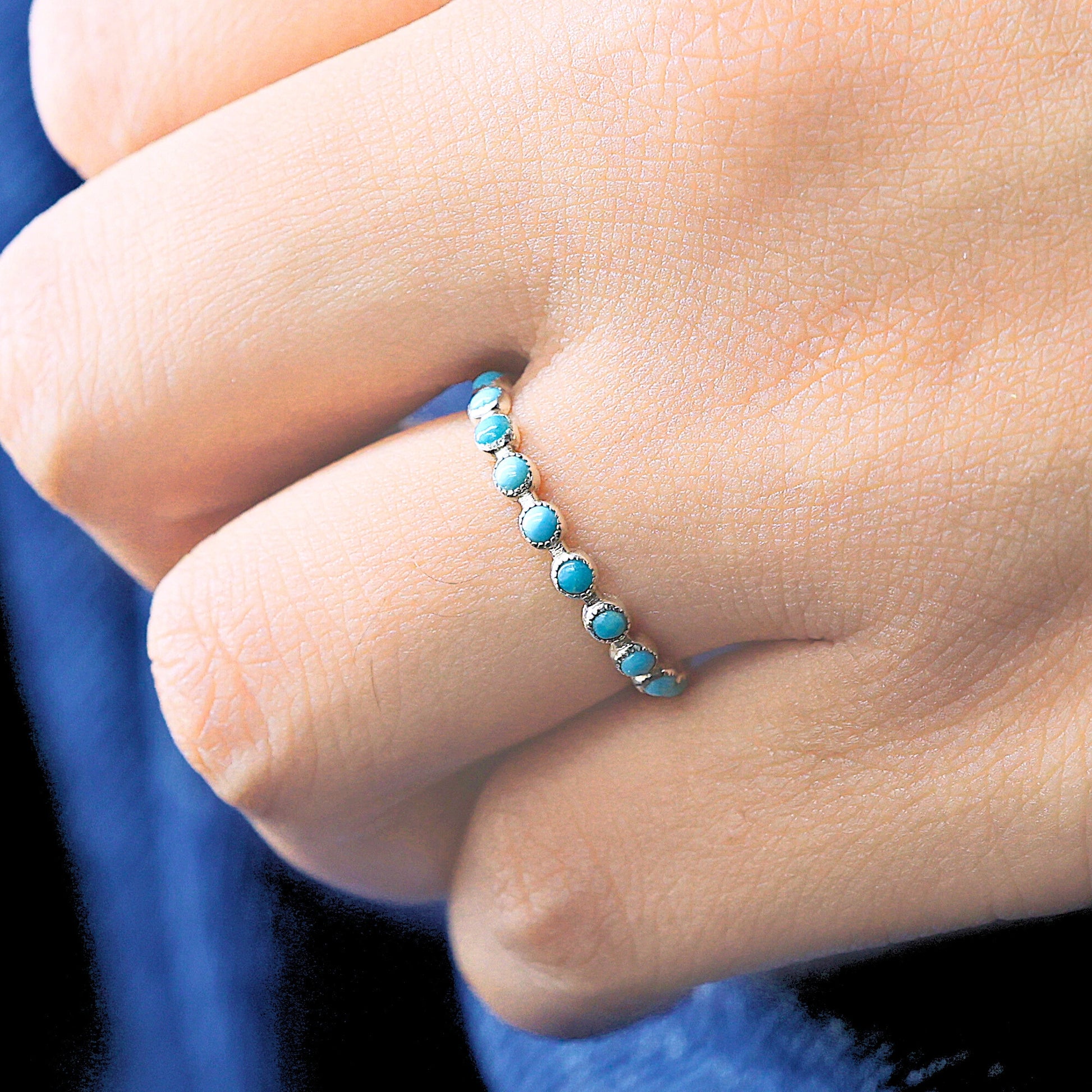 Close-up of a sterling silver turquoise ring eternity band with small blue stones on a hand over a dark blue fabric.