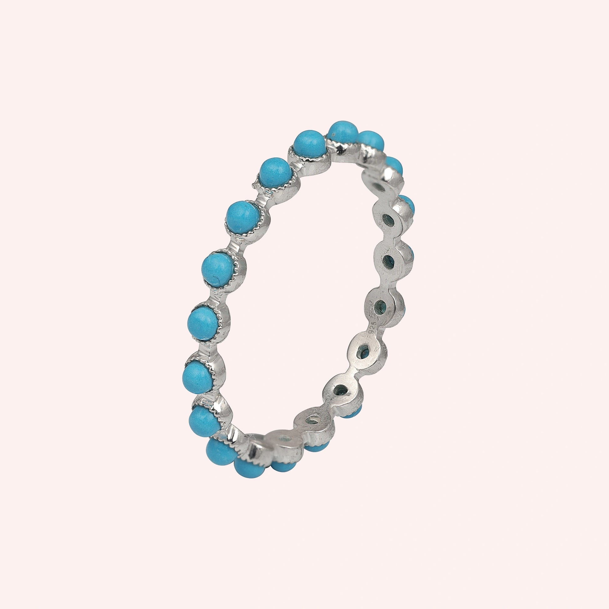 Angled sterling silver turquoise ring a full eternity band set with round blue turquoise stones on a smooth light pink background.